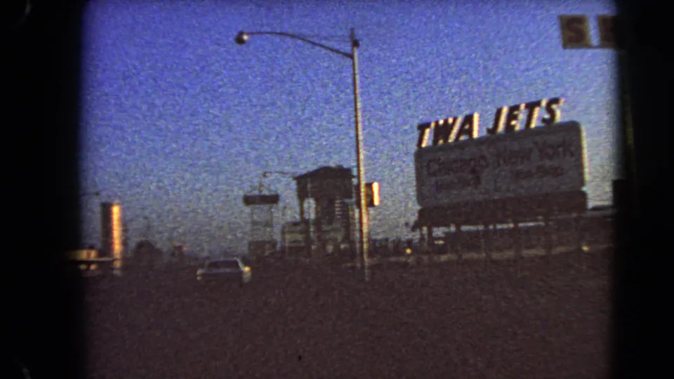 Drive-by 8mm frame in Las Vegas showing a large TWA Jets billboard near the road, with streetlights and a deep blue twilight sky.