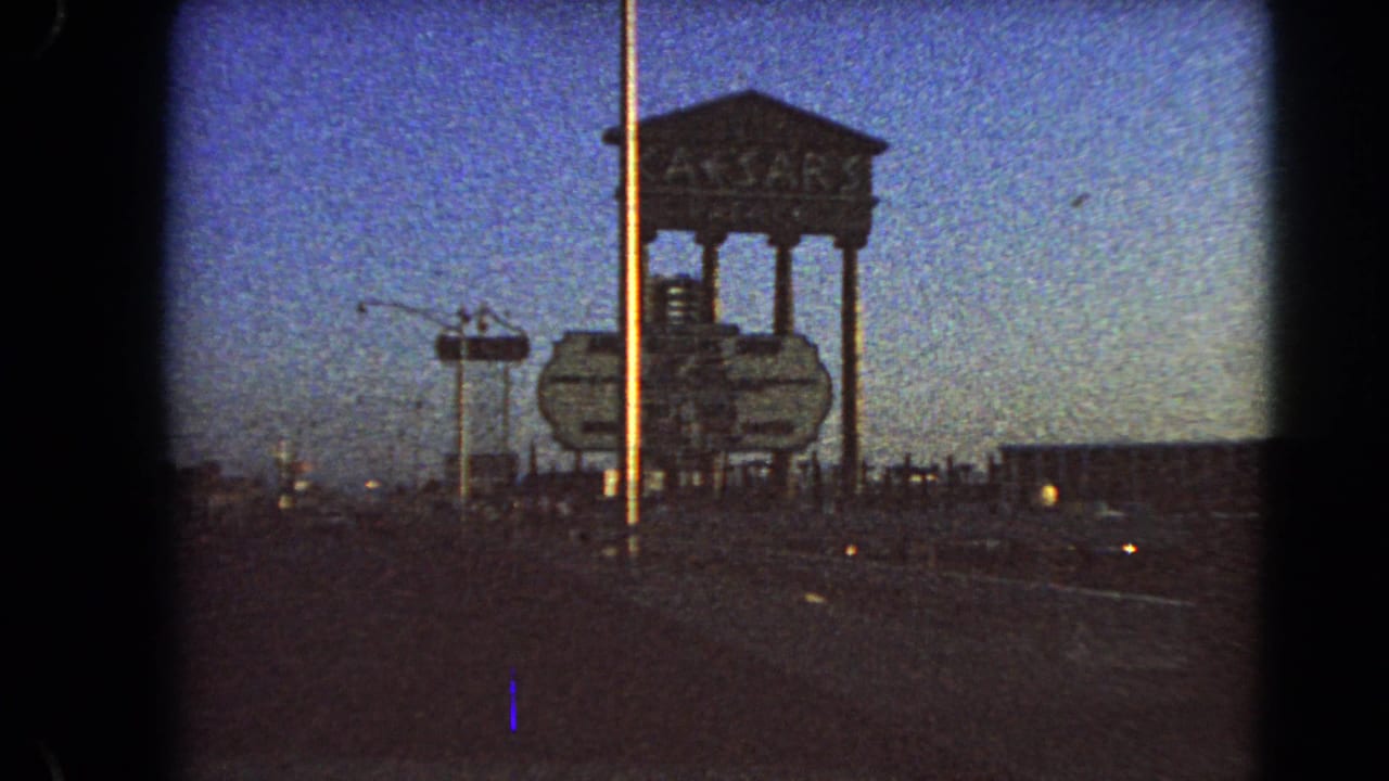 1966 Las Vegas roadside view showing the Caesars Palace sign structure, a tall pole, roadway, and dim evening sky from a moving car.
