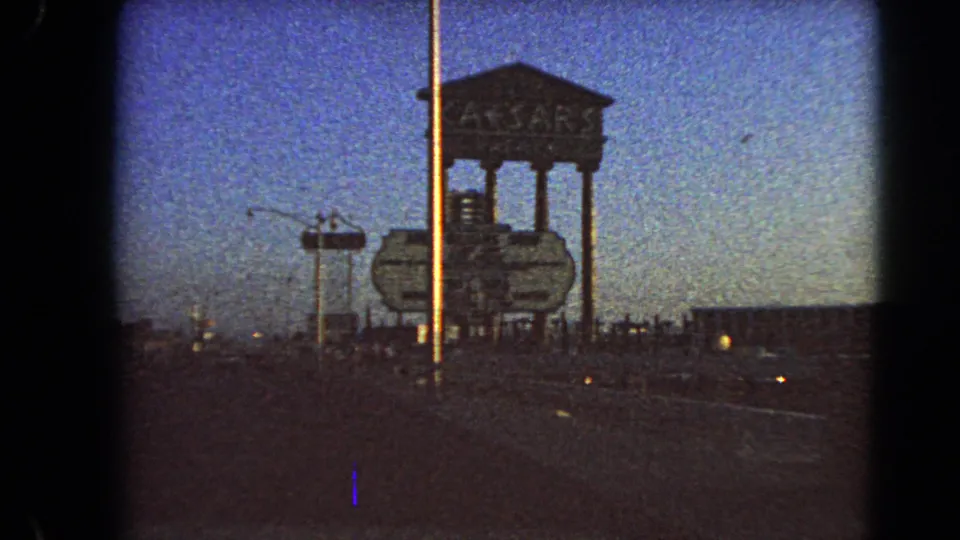 1966 Las Vegas roadside view showing the Caesars Palace sign structure, a tall pole, roadway, and dim evening sky from a moving car.