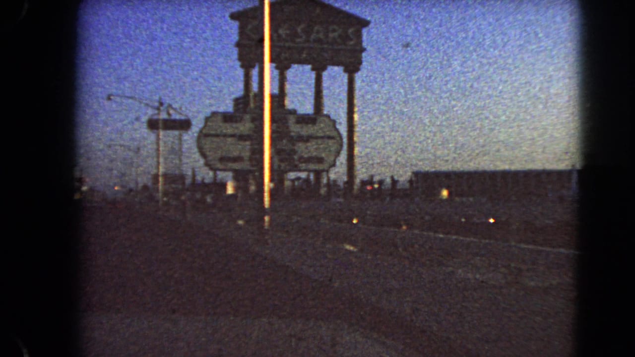 Las Vegas 8mm frame showing the Caesars Palace sign along a roadside at dusk, with streetlights, open road, and visible film grain.