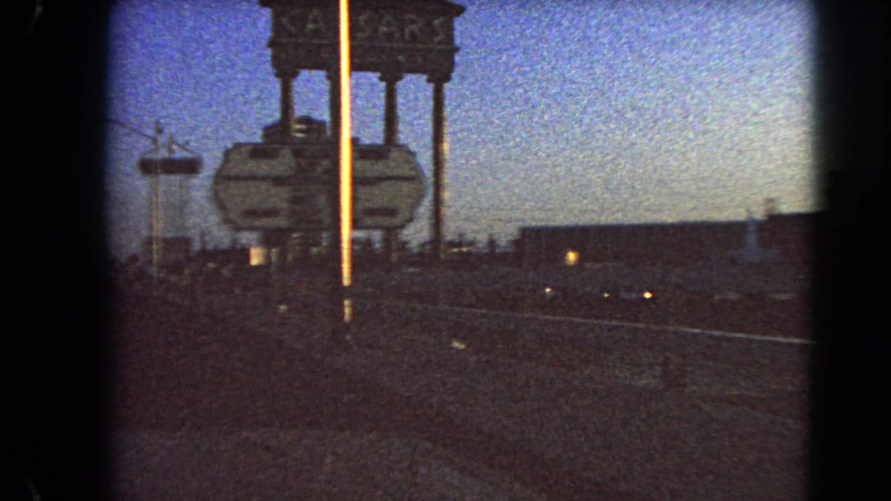 1966 Las Vegas 8mm frame showing the Caesars Palace sign structure beside a road at dusk, with cars and parking-lot lights below.