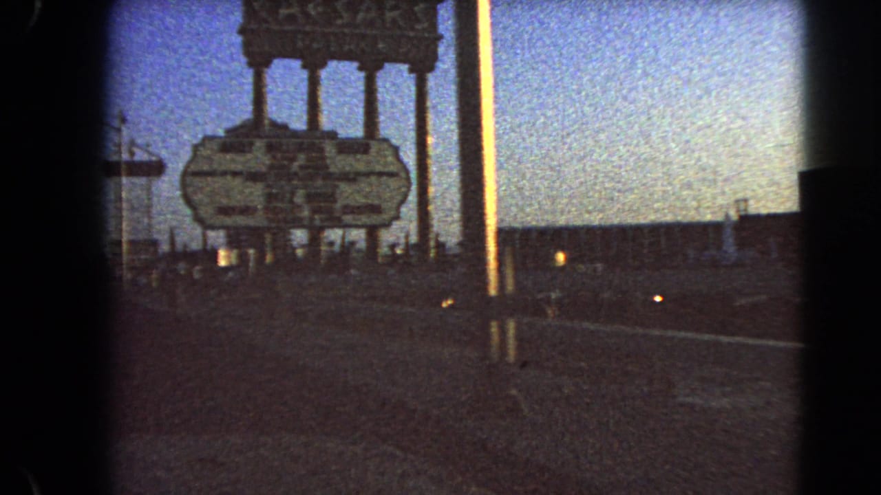 The Caesars Palace roadside sign appears against a blue dusk sky in Las Vegas, with a parking lot, cars, and small lights in the foreground.