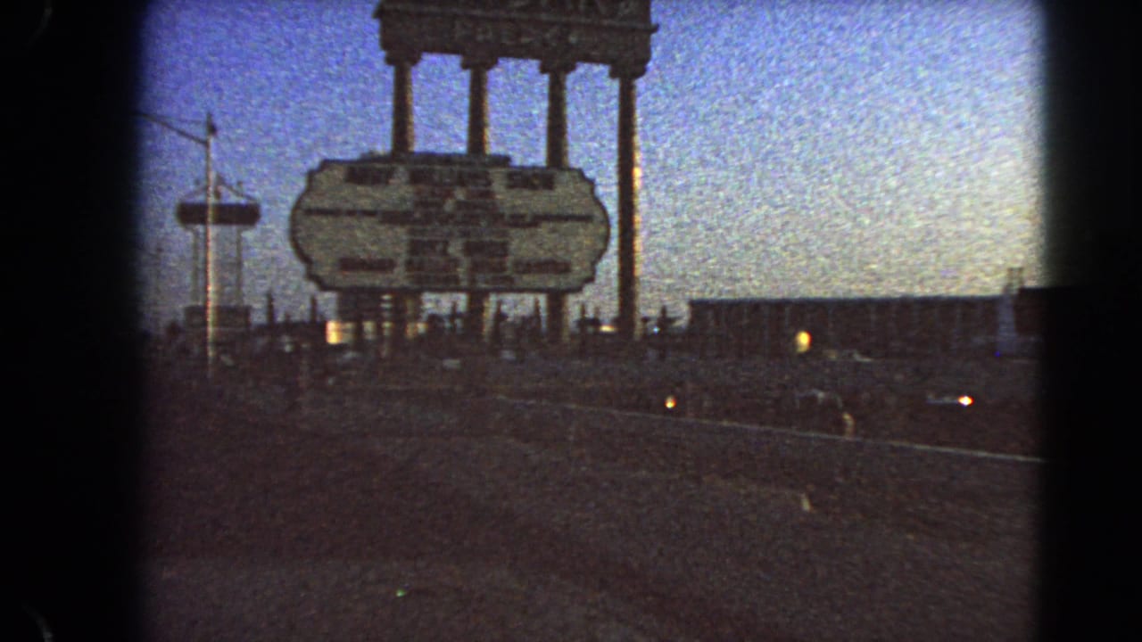 Centered 8mm view of the Caesars Palace sign in 1966 Las Vegas, with column supports, a directional board, road surface, and dim parking-lot lights.