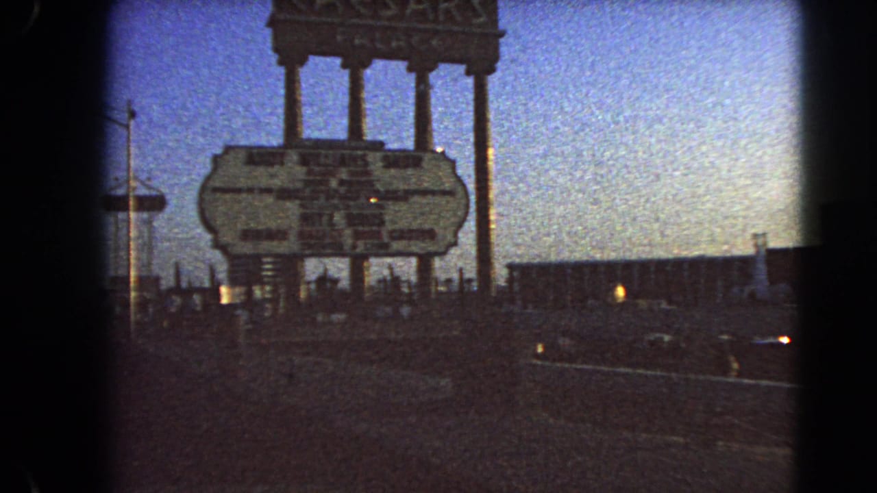 A 1966 Las Vegas home-movie frame shows the Caesars Palace sign from the road, with a dark parking area, scattered cars, and dusk sky.