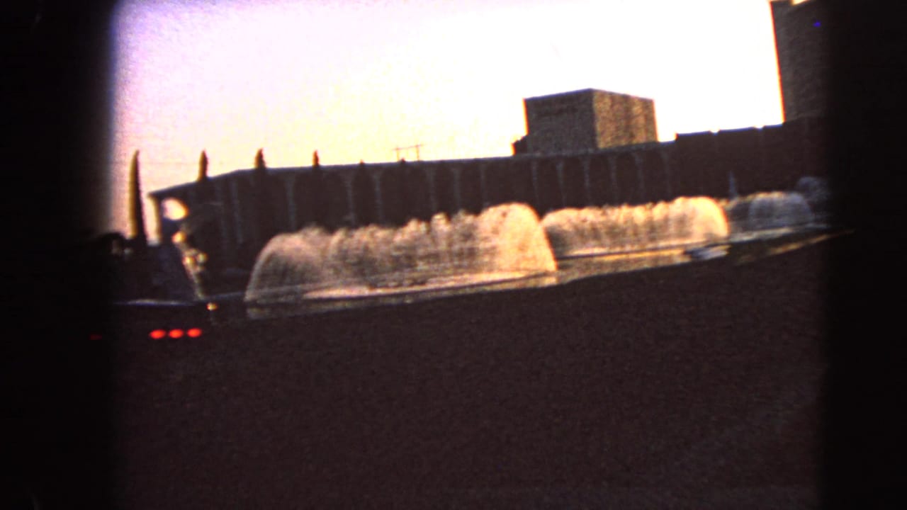 Fountain jets in front of dark building facades, seen from a low roadside angle.