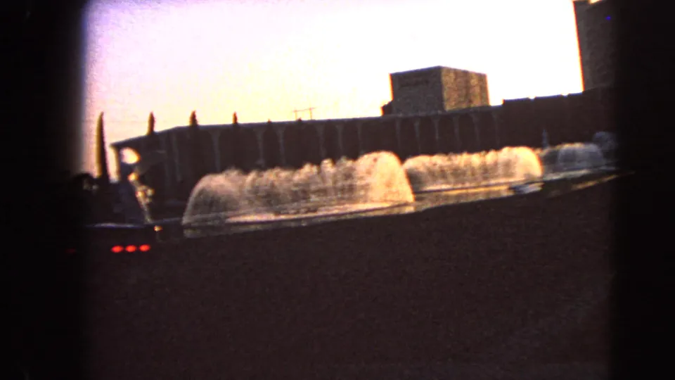 1966 Las Vegas home-movie frame showing illuminated fountain spray beside mid-century buildings from a moving roadside viewpoint.