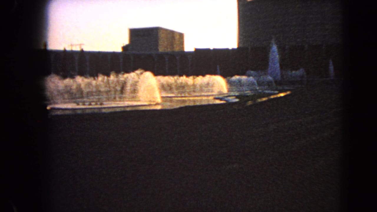 1966 Las Vegas 8mm frame showing several fountain plumes and long building facades, with dark vignette and roadside foreground.