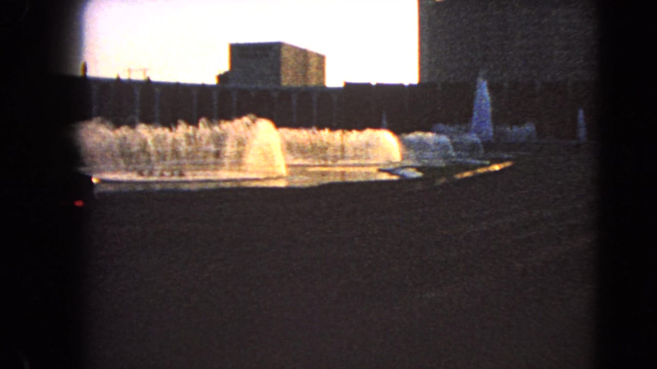 Fountain jets rise in front of dark modern buildings under a bright sky.