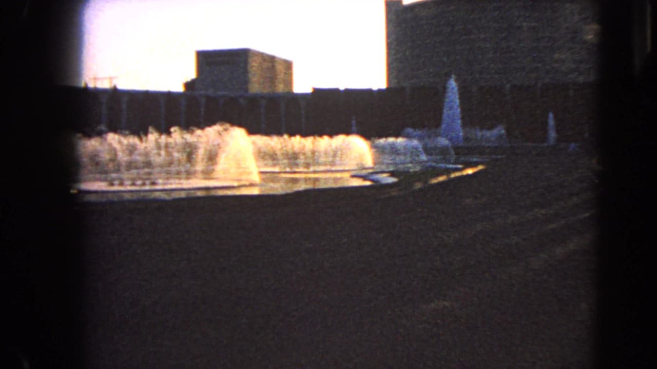 Wide fountain display with water spray, pool edge, and shaded buildings behind it.