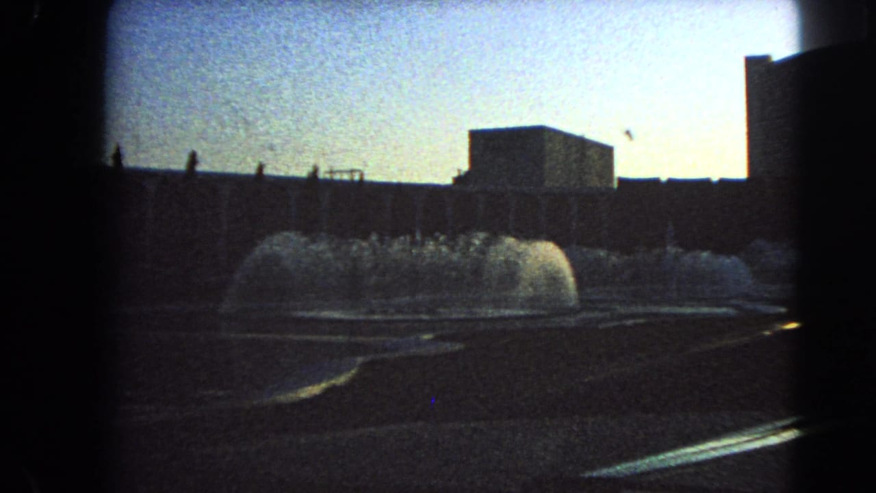 1966 Las Vegas 8mm still with a shadowed foreground, fountain spray, and rectangular modern building under bright daylight.