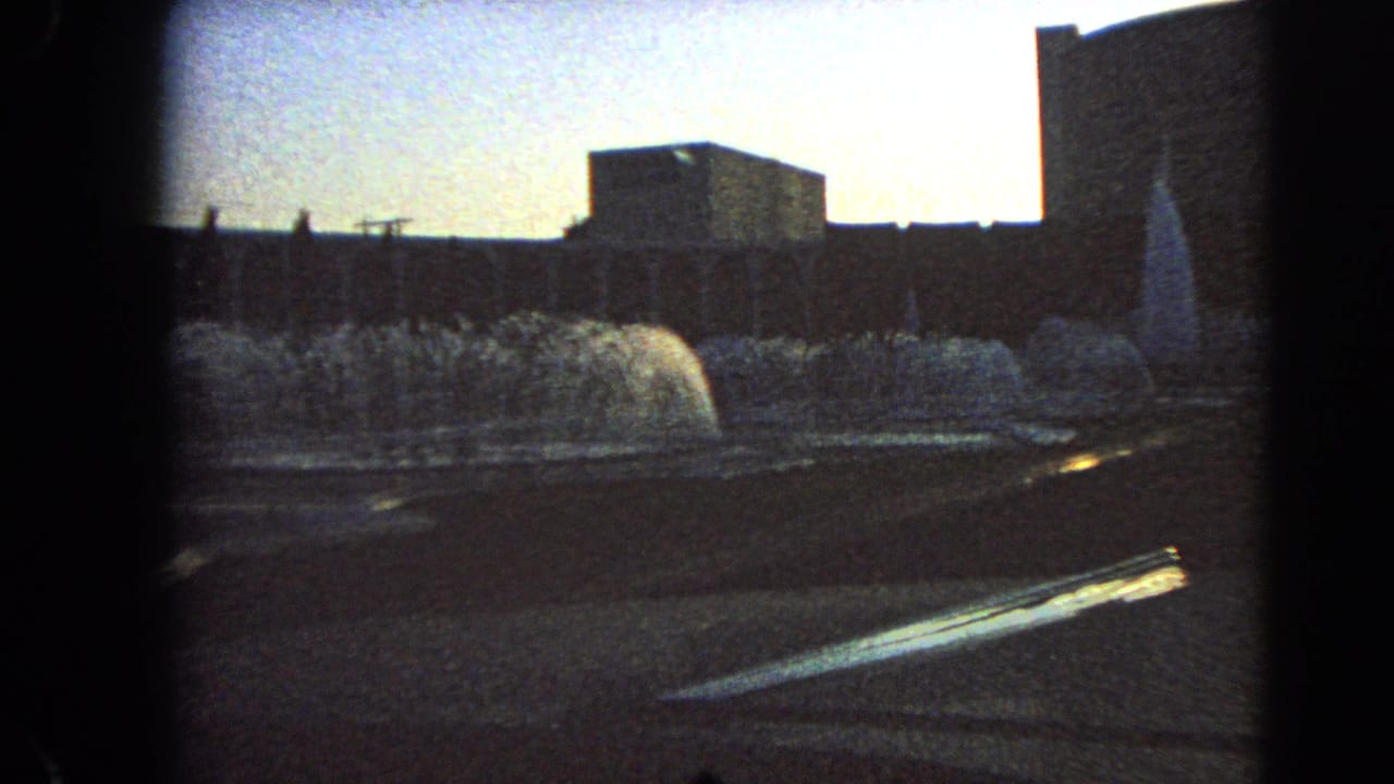 Final 1966 Las Vegas frame showing fountain jets, a paved edge, and dark modern building silhouettes with 8mm vignetting.
