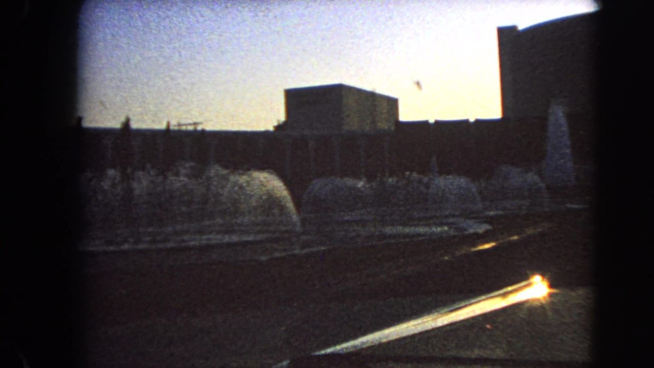 Moving-car view in Las Vegas, 1966, with fountain arcs, silhouetted buildings, and sun flare on 8mm film.