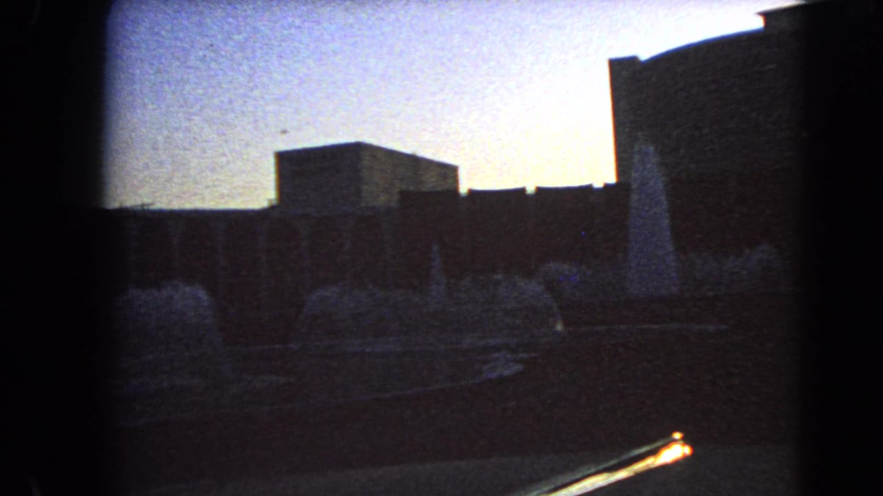 Low-light 8mm view of a Las Vegas courtyard fountain at dusk, framed by dark building masses and visible film-edge shadow.
