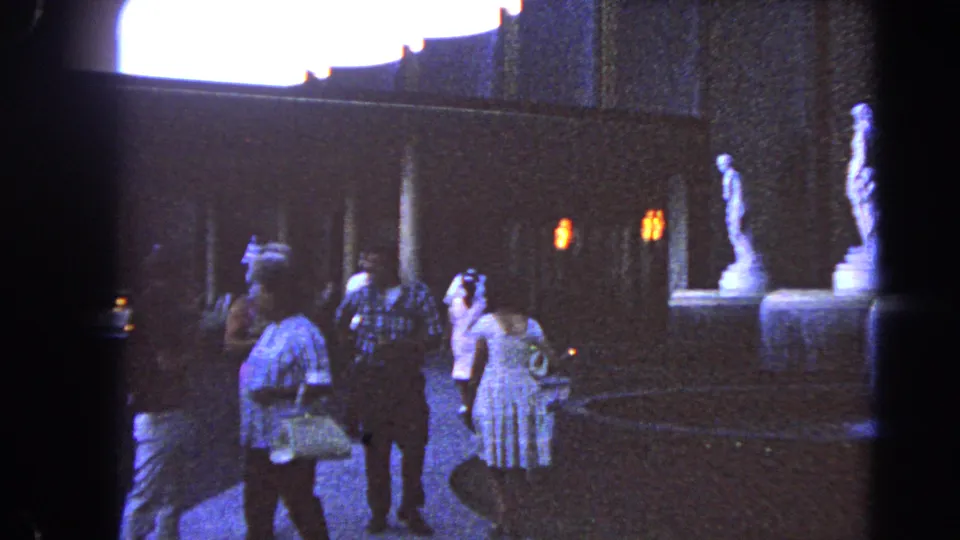 Handheld 8mm frame of Las Vegas visitors strolling through an evening courtyard with statues, paving, and warm small lights.