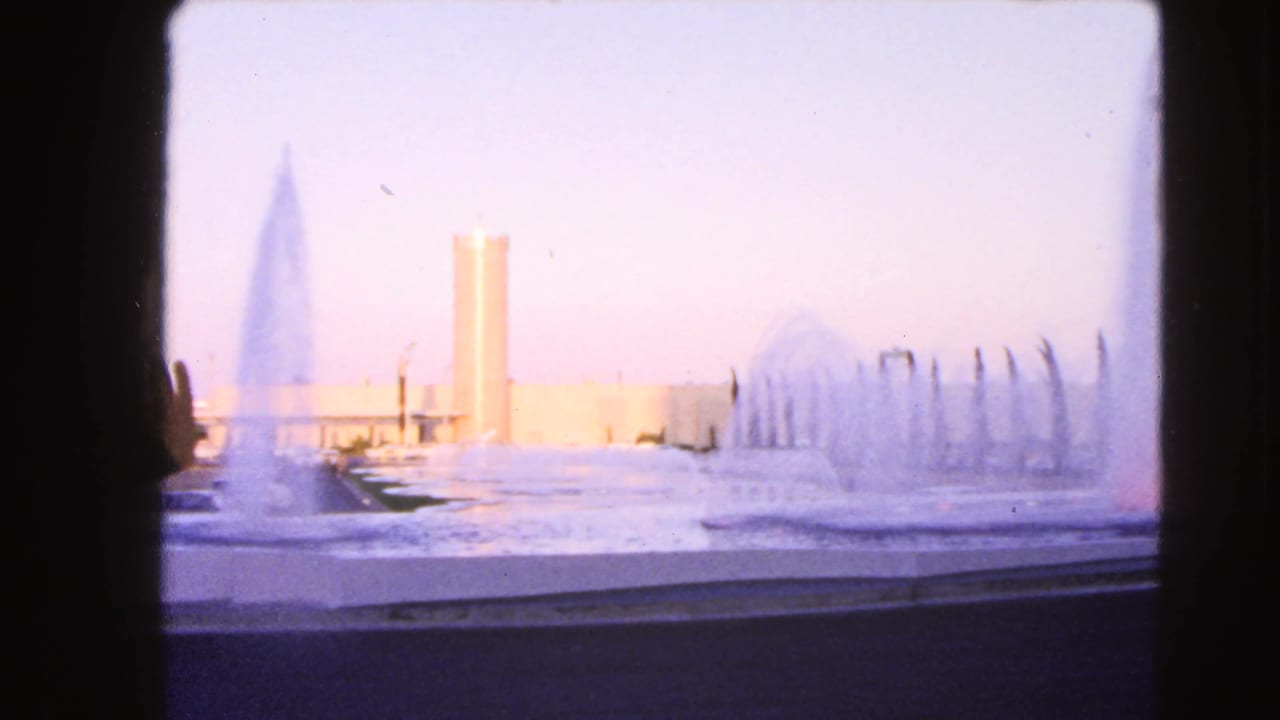 1966 Las Vegas home-movie frame showing a soft, overexposed fountain scene with water jets and a tall cylindrical tower in the distance.