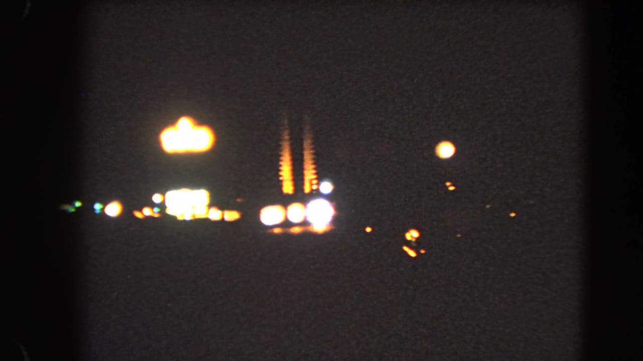 Soft 1966 Las Vegas night frame showing a bright distant sign shape, roadside lights, and deep underexposed 8mm texture.