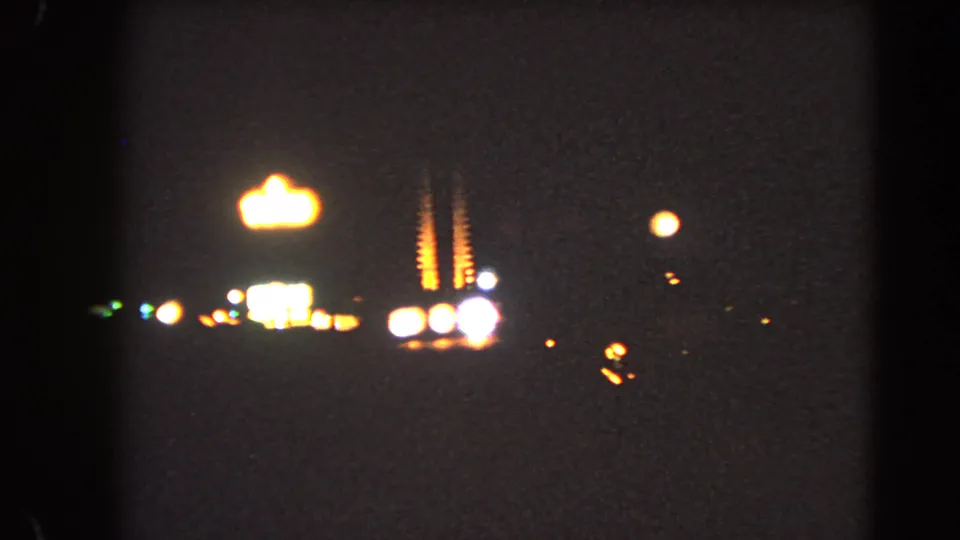 Soft 1966 Las Vegas night frame showing a bright distant sign shape, roadside lights, and deep underexposed 8mm texture.