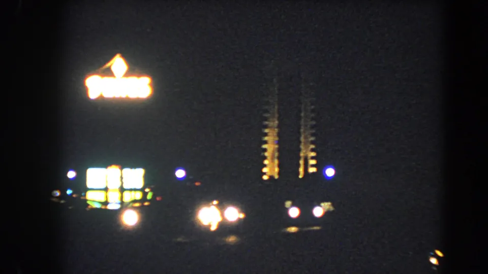 1966 Las Vegas Strip night view with blurred illuminated signage, neon columns, car headlights, and dense 8mm grain.