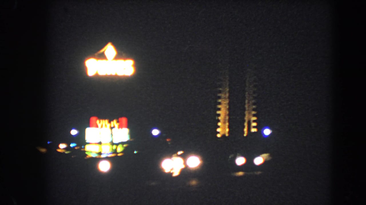 1966 Las Vegas night frame with glowing roadside neon, dark sky, and passing vehicle headlights on 8mm film.