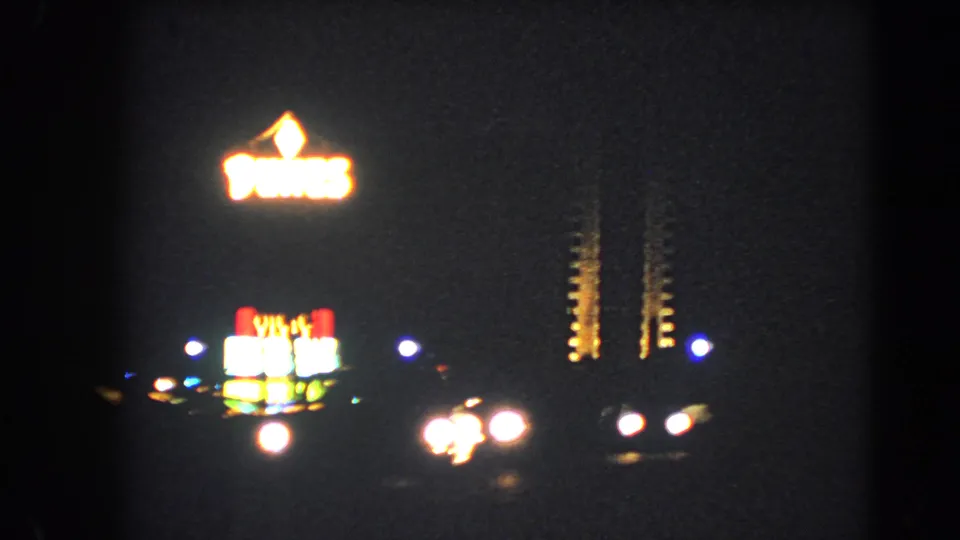 1966 Las Vegas night frame with glowing roadside neon, dark sky, and passing vehicle headlights on 8mm film.