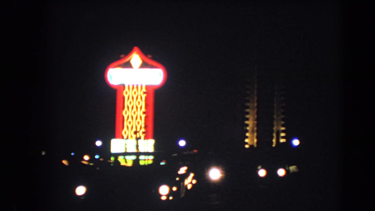 Tall illuminated Las Vegas sign at night, filmed in 1966 with blooming neon and roadside car lights.