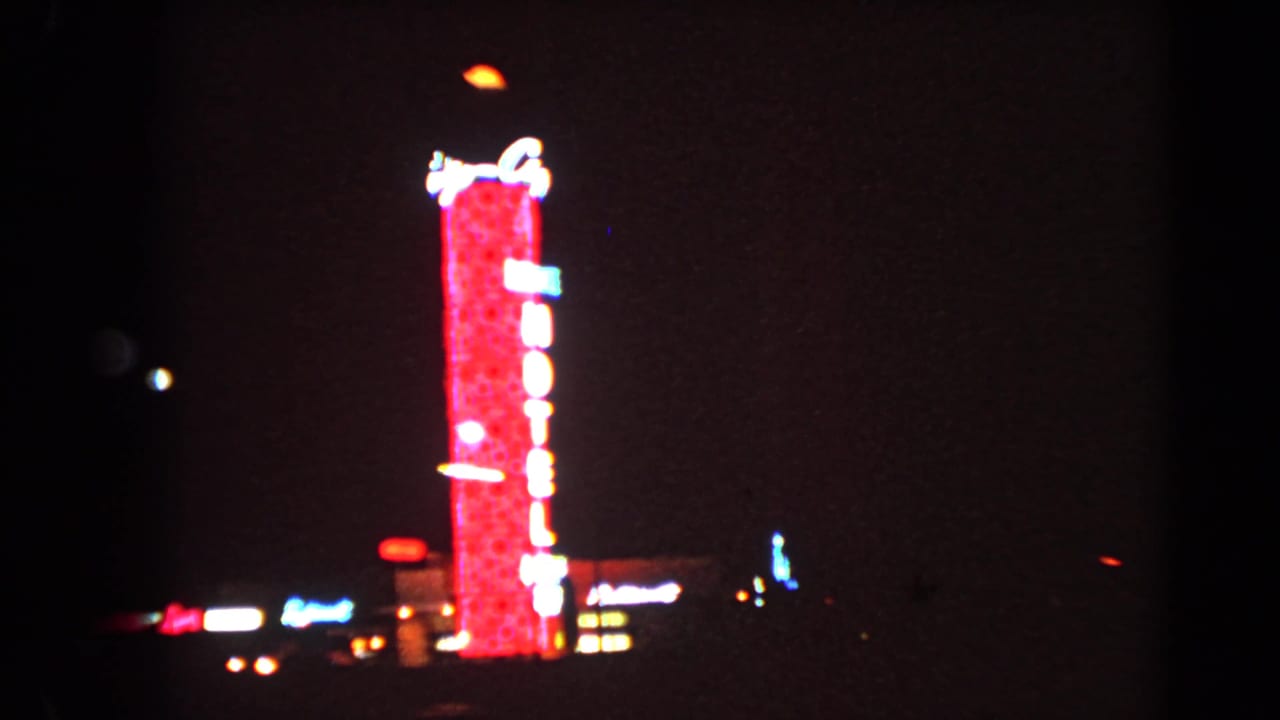 1966 Las Vegas nighttime frame with a red vertical hotel neon sign and scattered street lights in the distance.
