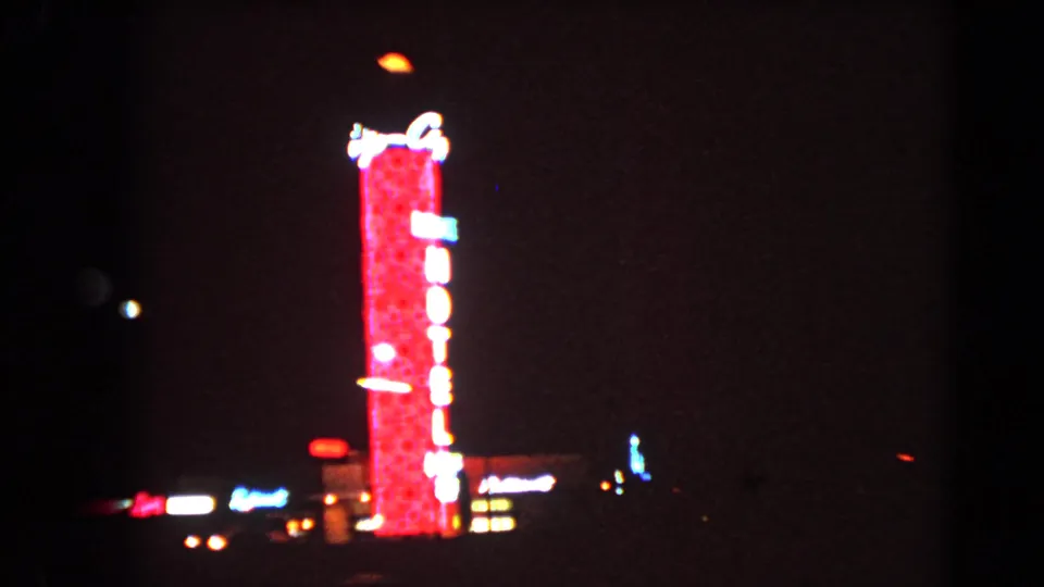 1966 Las Vegas nighttime frame with a red vertical hotel neon sign and scattered street lights in the distance.