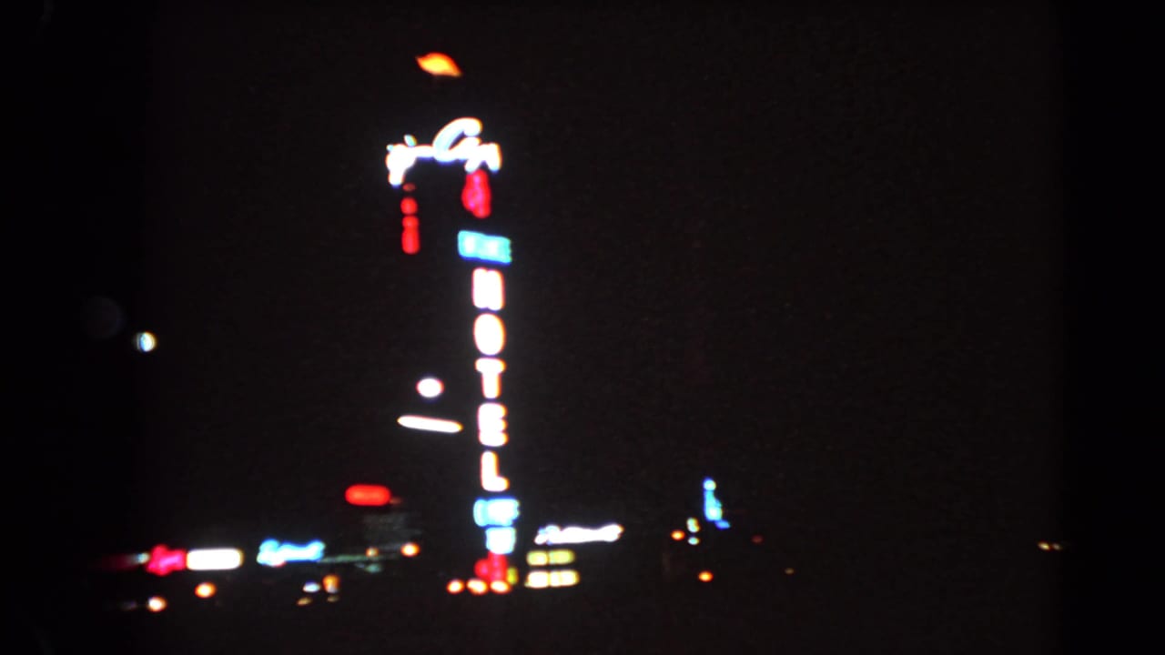 1966 Las Vegas 8mm frame showing a tall illuminated hotel sign and scattered city lights at night.