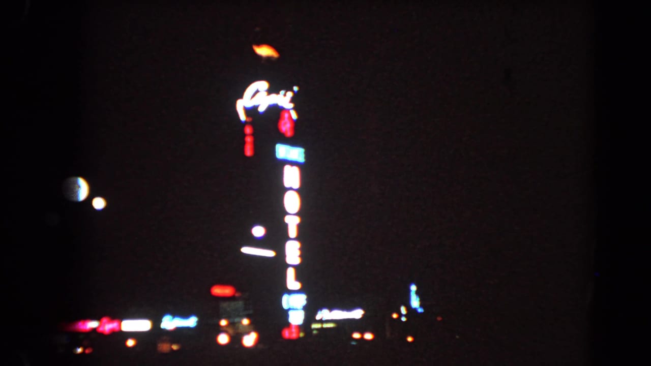 1966 Las Vegas night frame with a glowing vertical hotel sign, dark sky, and distant illuminated signage.