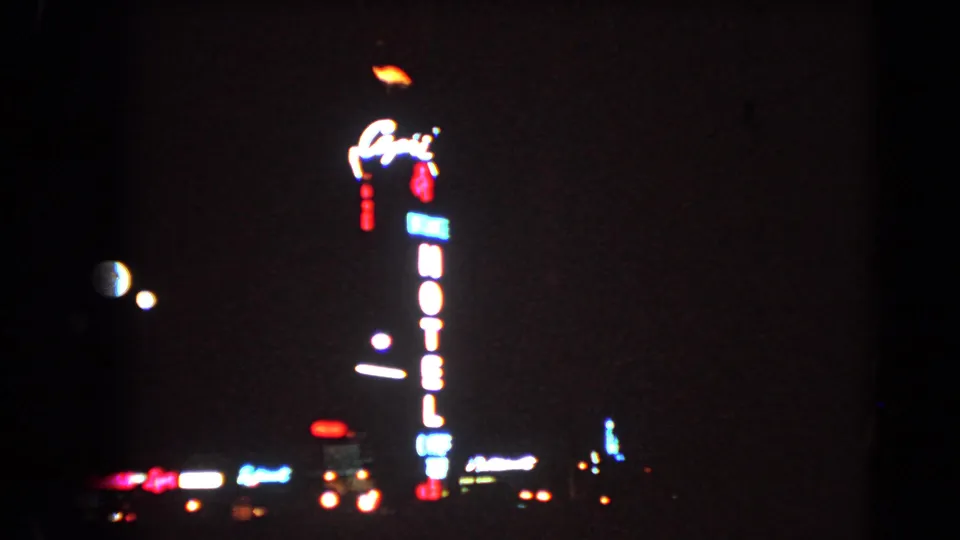 1966 Las Vegas night frame with a glowing vertical hotel sign, dark sky, and distant illuminated signage.