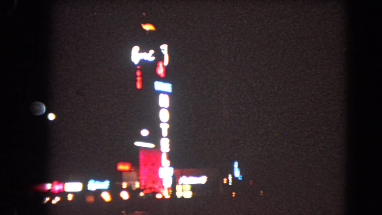 1966 Las Vegas 8mm frame with blooming neon, a vertical hotel sign, and scattered lights near the street line.