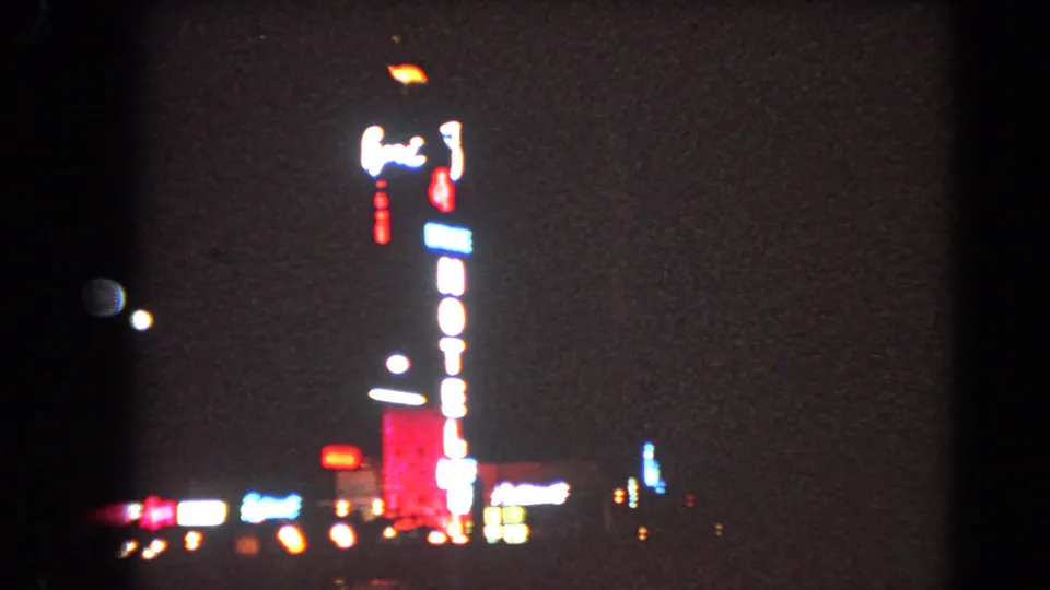 1966 Las Vegas 8mm frame with blooming neon, a vertical hotel sign, and scattered lights near the street line.