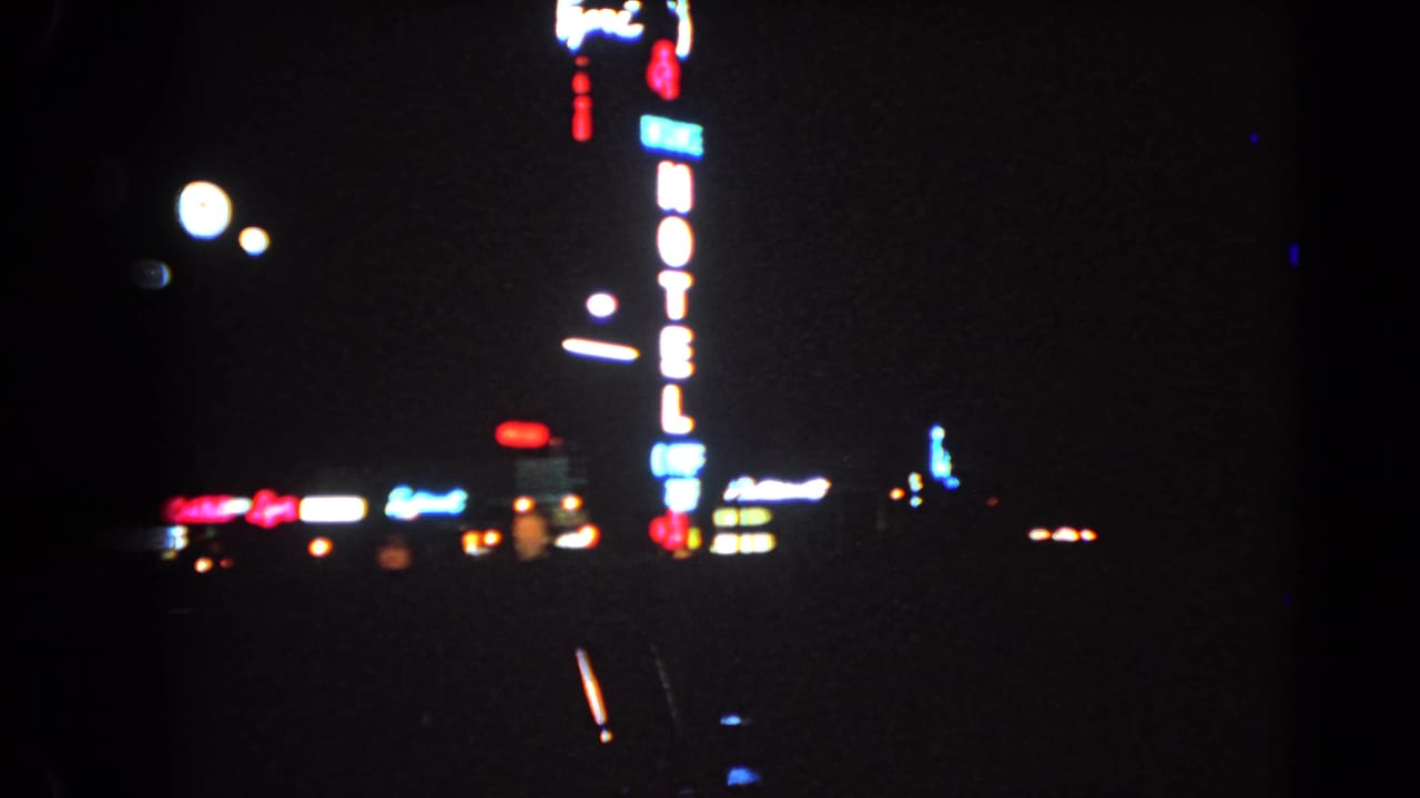 A moving 8mm view of Las Vegas at night shows a vertical neon hotel sign, blurred lights, and dark road foreground.
