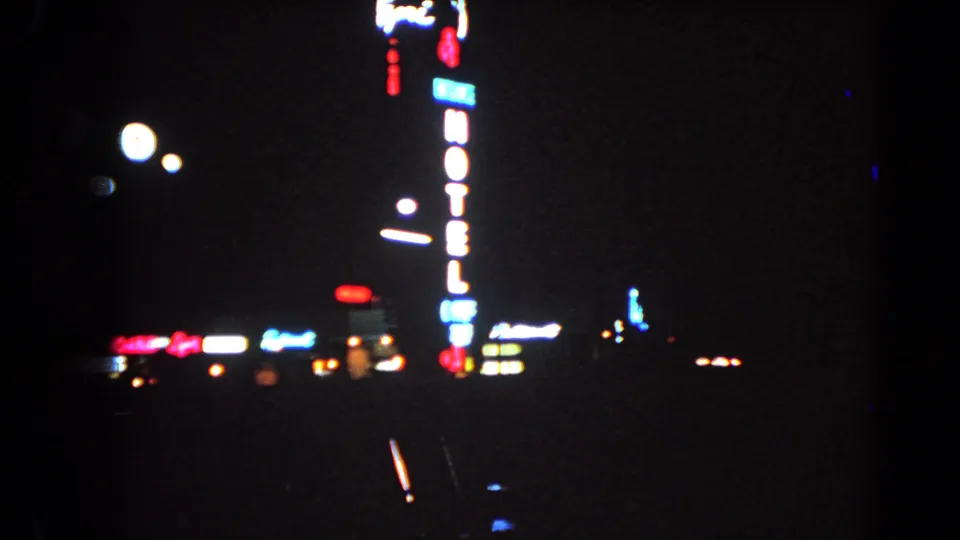 A moving 8mm view of Las Vegas at night shows a vertical neon hotel sign, blurred lights, and dark road foreground.
