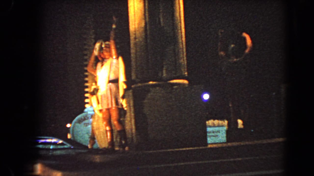 1966 Las Vegas street-level frame shows a person in a light dress near a lit column, with dark building frontage and roadway nearby.