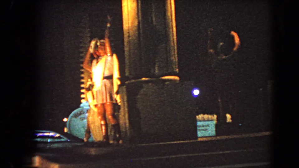 1966 Las Vegas street-level frame shows a person in a light dress near a lit column, with dark building frontage and roadway nearby.
