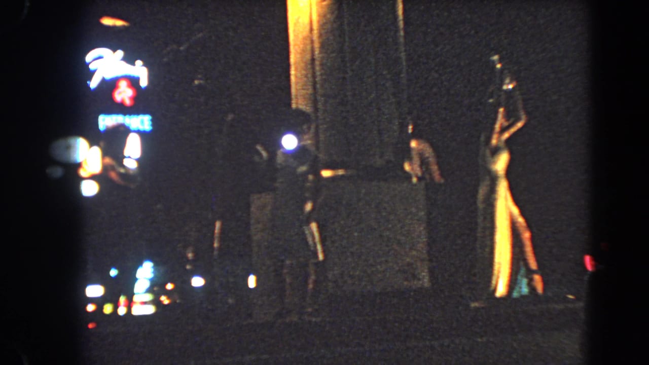 1966 Las Vegas night footage shows dressed women near a sidewalk entrance with neon signs and traffic lights in the distance.