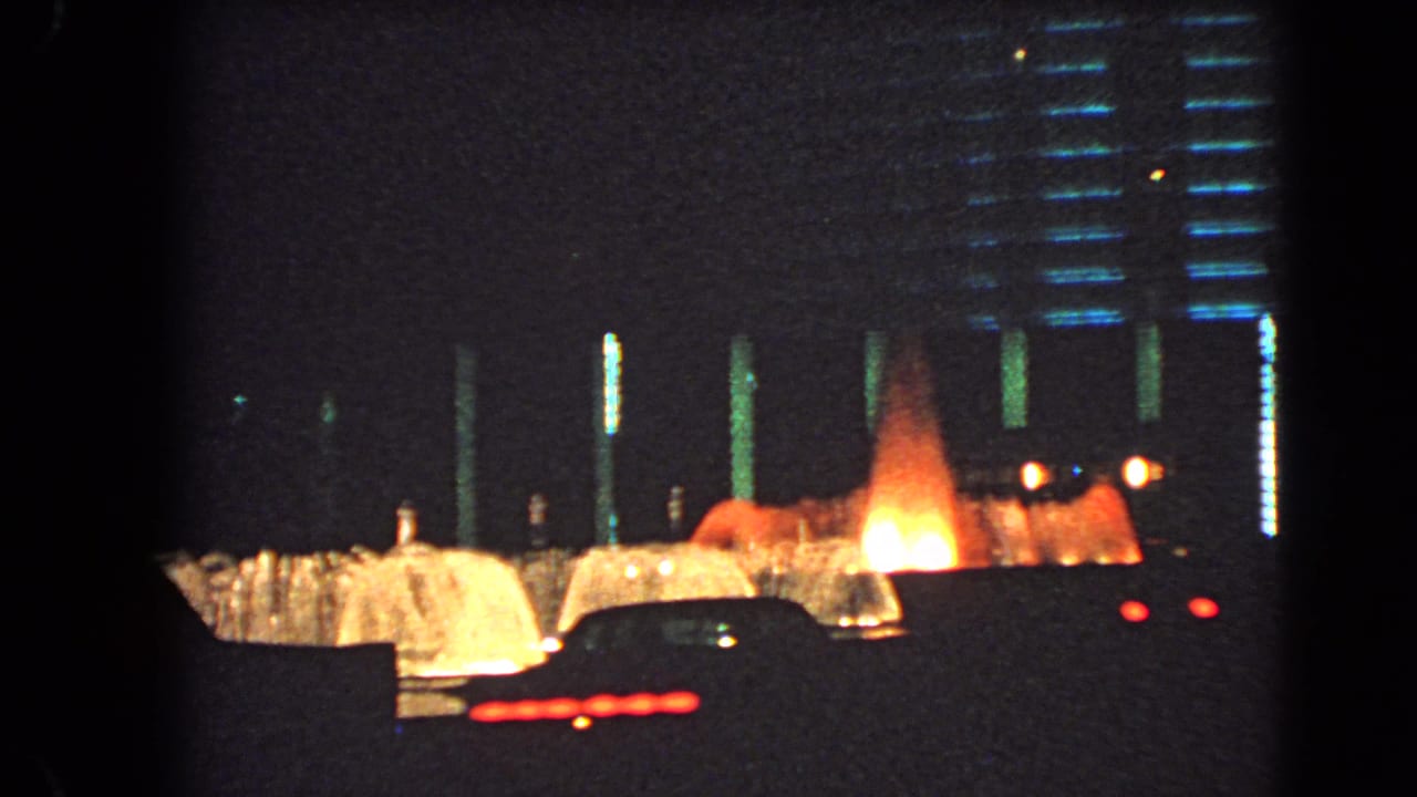 A night view of Las Vegas fountains shows warm water jets behind silhouetted cars and lit building details.