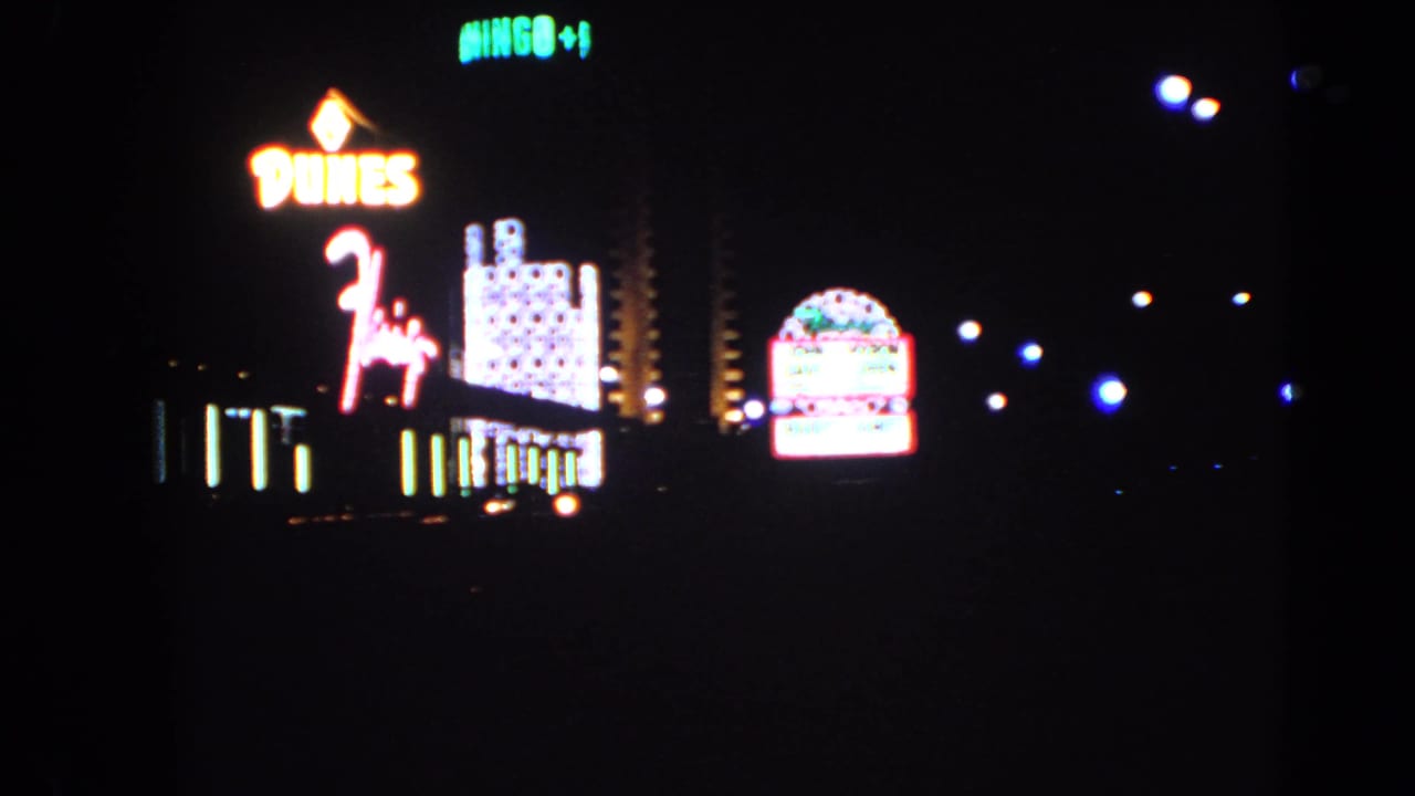1966 Las Vegas home-movie frame showing Dunes neon, a lit hotel tower, vertical sign lights, and a bright roadside marquee.