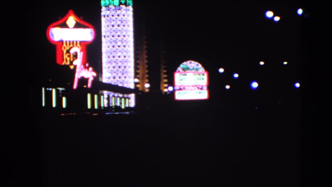 1966 Las Vegas frame with a bright casino marquee, Dunes-style horse-and-rider neon, tall lit tower, and distant street lamps at night.