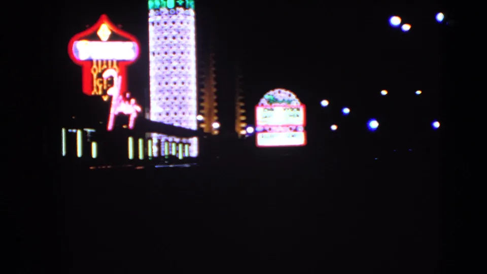 1966 Las Vegas frame with a bright casino marquee, Dunes-style horse-and-rider neon, tall lit tower, and distant street lamps at night.