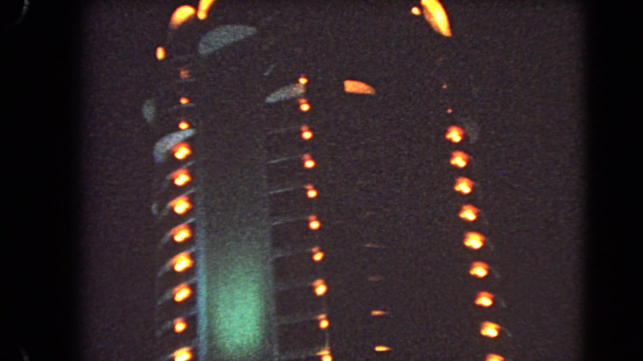 1966 Las Vegas 8mm frame showing a closer, soft-focus facade with orange light rows, green illumination, and visible film texture.