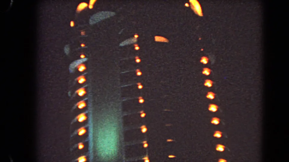 1966 Las Vegas 8mm frame showing a closer, soft-focus facade with orange light rows, green illumination, and visible film texture.