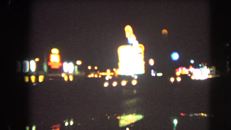 1966 Las Vegas night-driving frame with distant neon signs, bright roadside advertising, and car headlights blurred by motion and low light.