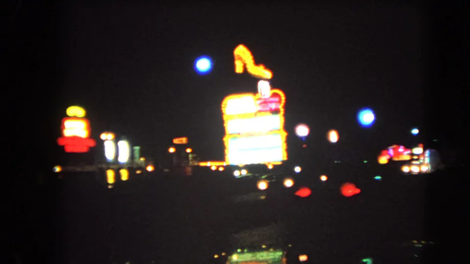 1966 Las Vegas night frame showing a luminous roadside sign topped by a high-heeled shoe shape, with small traffic lights and neon dots in the distance.