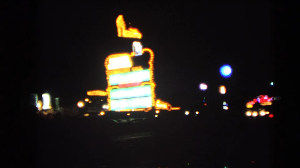 Closer 1966 Las Vegas night-driving frame with a high-heeled shoe sign, stacked neon panels, and distant roadside lights softened by handheld motion.
