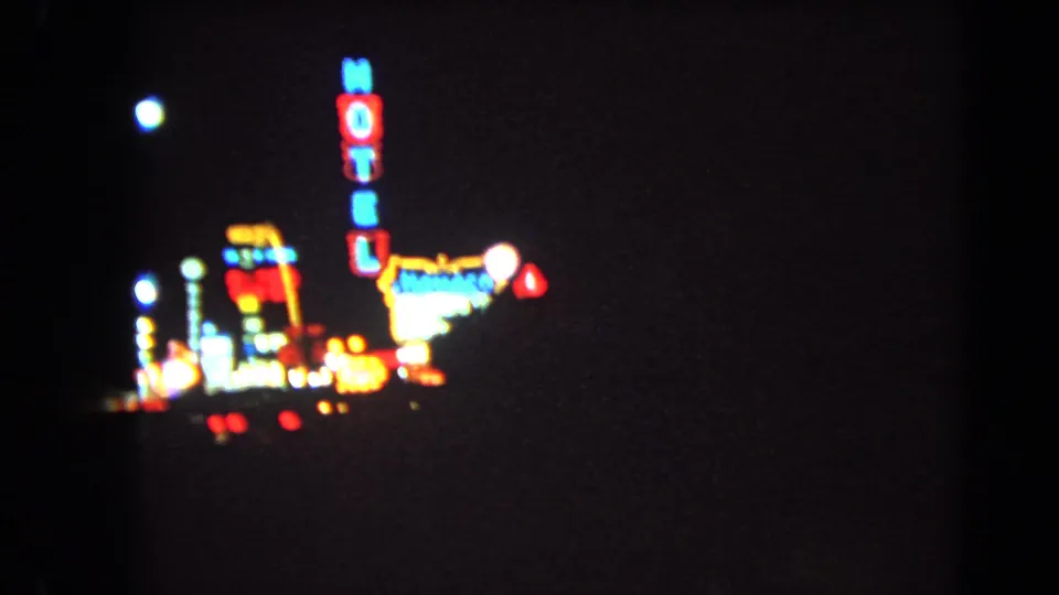 1966 Las Vegas night-driving frame with a vertical motel sign, clustered roadside neon, and red vehicle lights against a dark street scene.