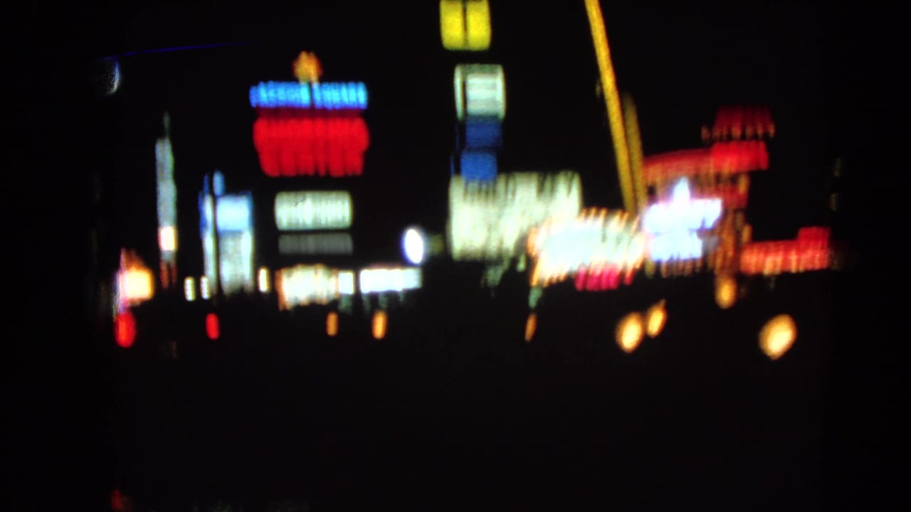 1966 Las Vegas night-drive frame with soft, blurred neon signs and roadside lights against a dark sky.