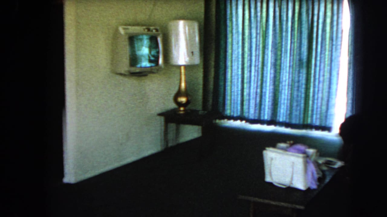 Dim hotel room with striped curtains, wall television, lamp, table, and white handbag.