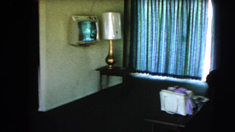 1966 Las Vegas hotel room interior with striped curtains, wall-mounted television, lamp, table, and white handbag visible.